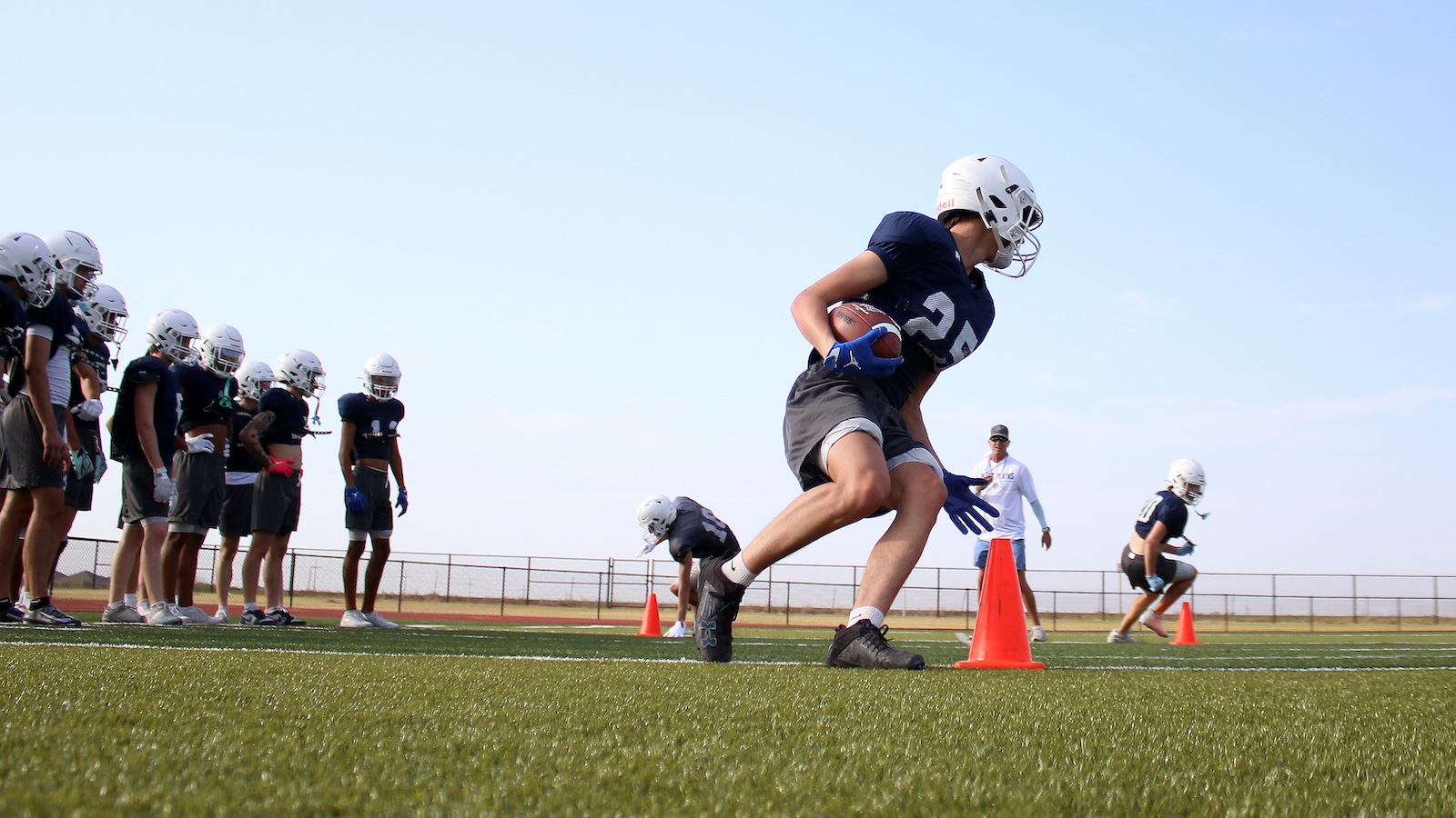 Football practice on a sunny field: players in navy jerseys lined up as a ballcarrier dashes past orange cones far right
