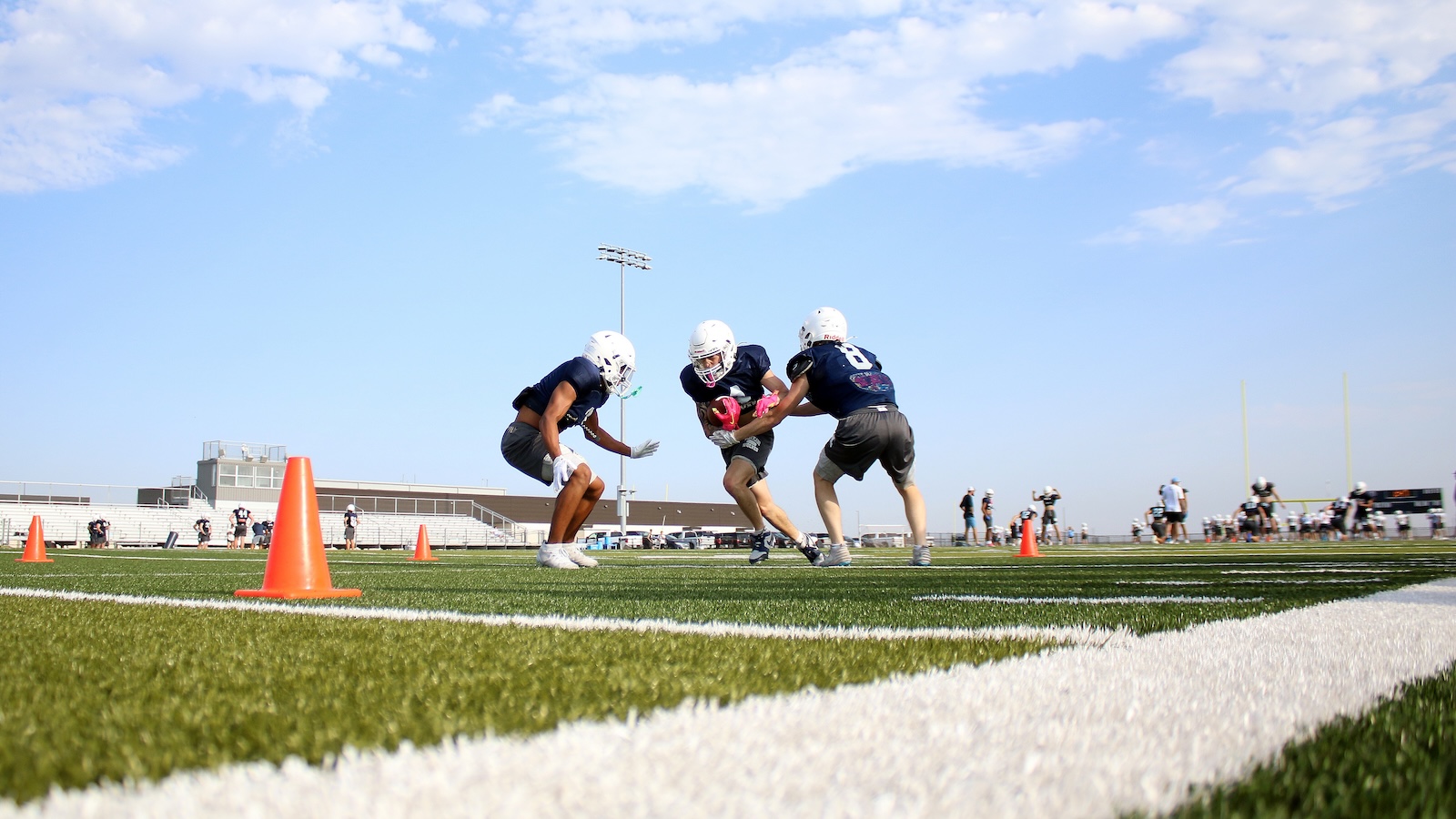 Three football players in navy uniforms rush a play on a green turf field dotted with orange cones under a clear blue sky.