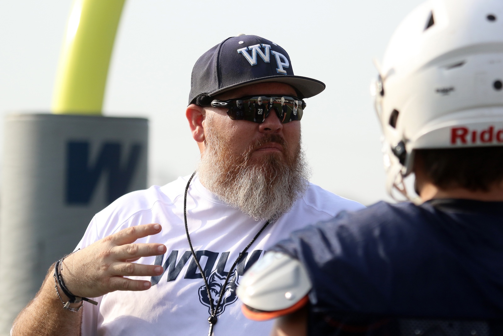 Bearded man in a dark cap with 'WP' and sunglasses speaks to a football player wearing a helmet in an outdoor setting.