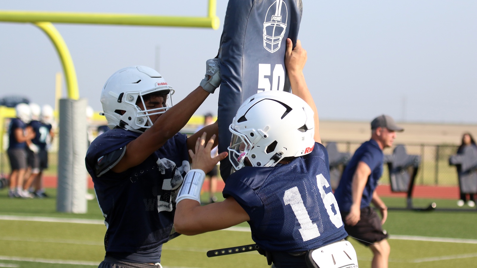 Two football players in navy jerseys and white helmets practice blocking a blue tackling shield on a sunny field.