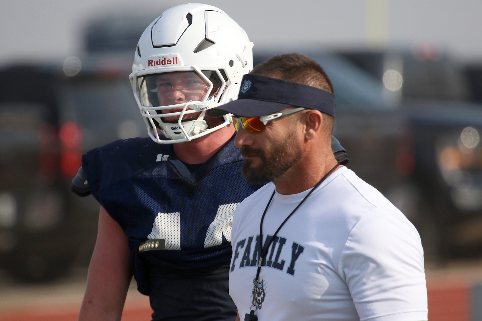 Football coach in a white 'FAMILY' shirt with a whistle stands beside a player in a white helmet and navy jersey on the field.