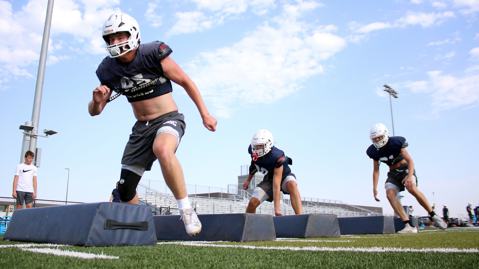 American football players sprint through a drill over padded blocking shields on a sunny turf field during practice, wearing helmets and navy uniforms.