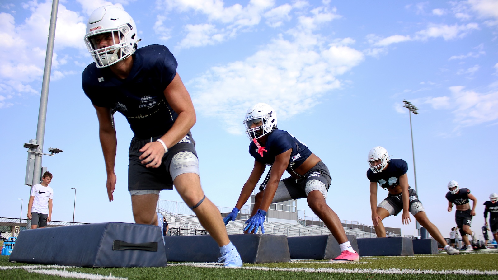 Football players in navy jerseys and white helmets sprint through padded obstacles during an agility drill on a sunny day.