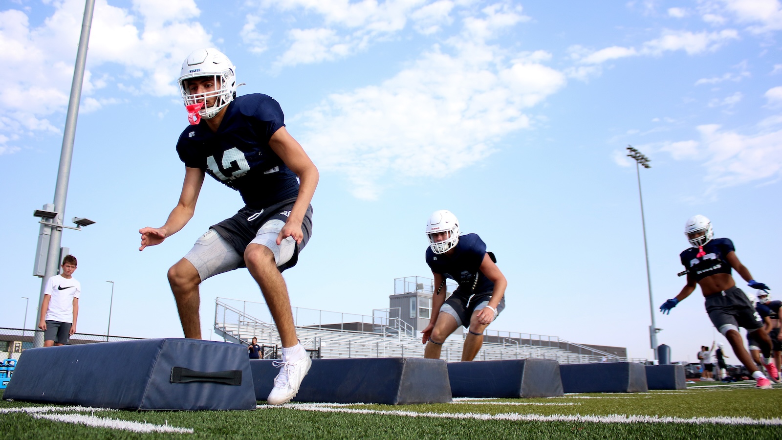 Football players in navy jerseys sprint over padded obstacles during practice on a sunny day, helmets on and bleachers in the background.