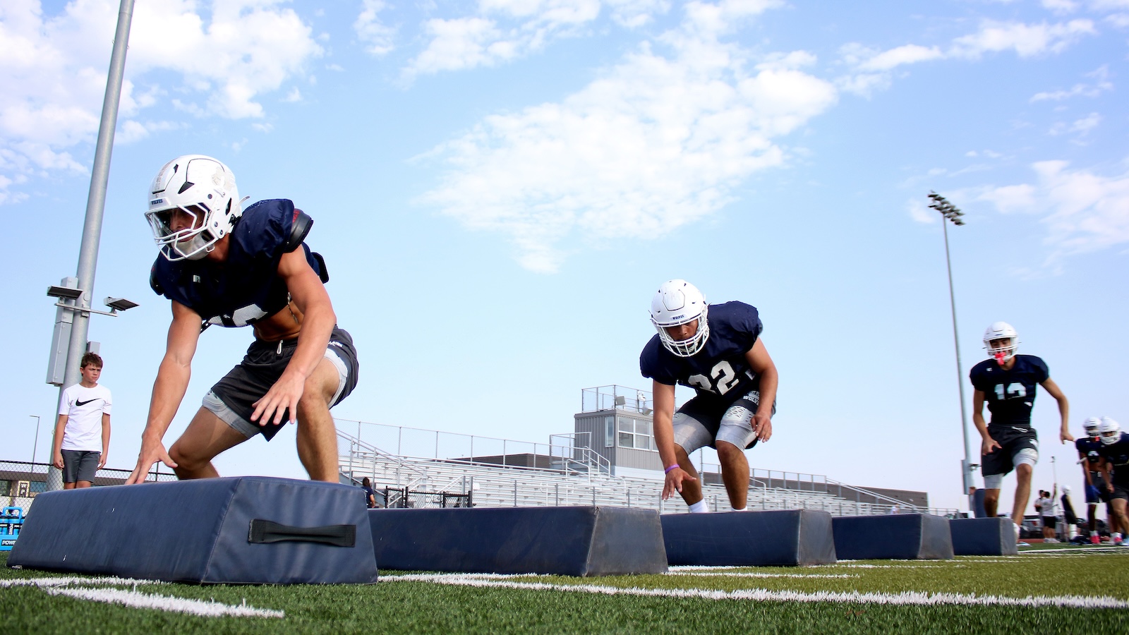 Football players in navy jerseys and white helmets sprint from starting pads on a green turf field during practice on a sunny day.