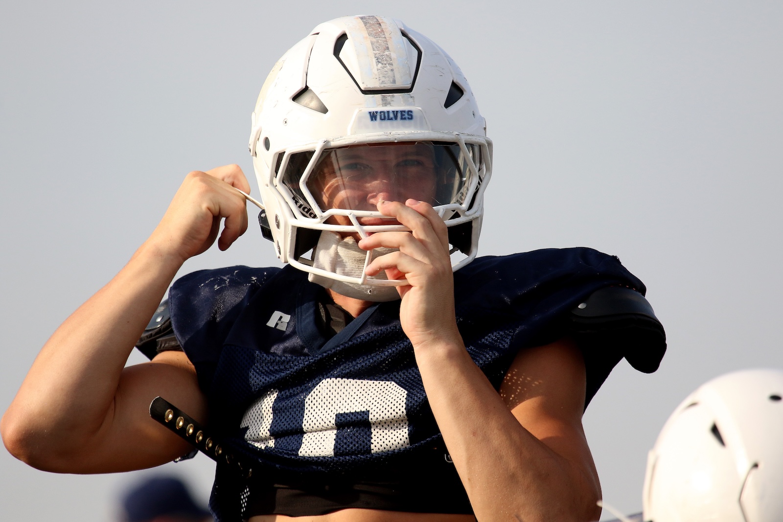 Football player in a navy jersey puts on a white helmet labeled WOLVES, adjusting the strap.