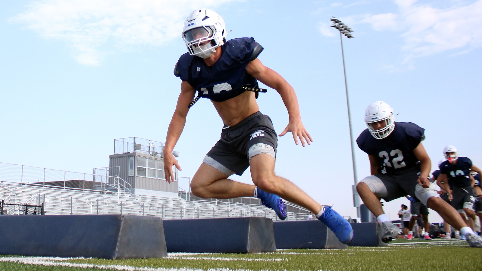 Football player in a navy jersey and white helmet jumps over padded barriers during a practice drill on a football field, other players in the background.