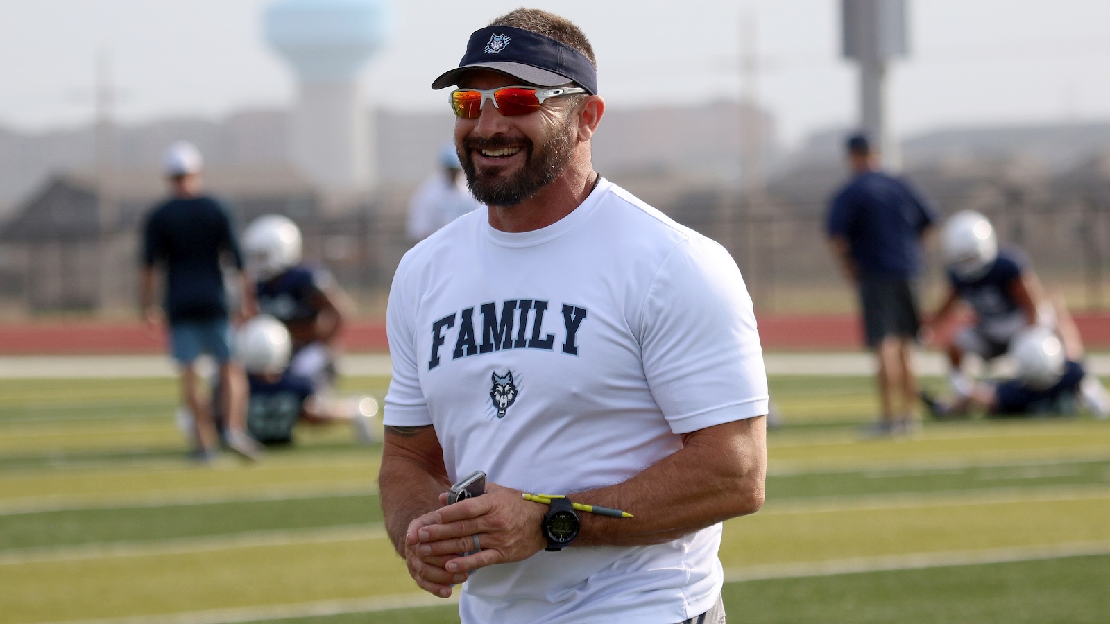 Smiling man in a white 'FAMILY' shirt and navy visor on a football practice field, with players in the background.