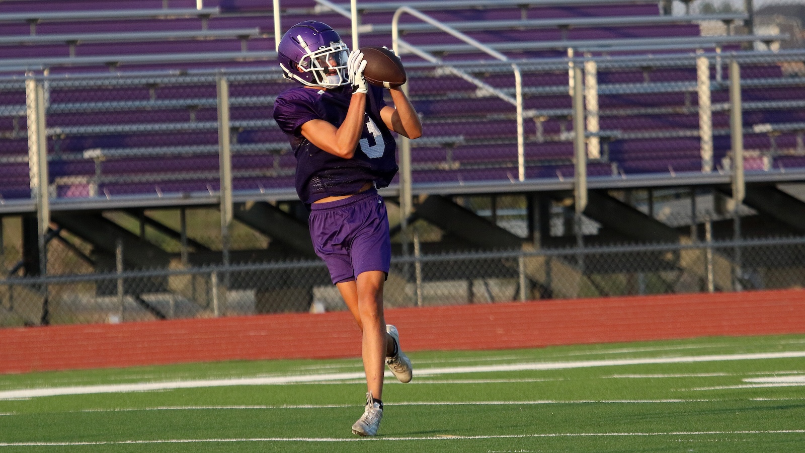 Football player in a purple uniform catches a football midair on a green turf field with purple bleachers behind him.
