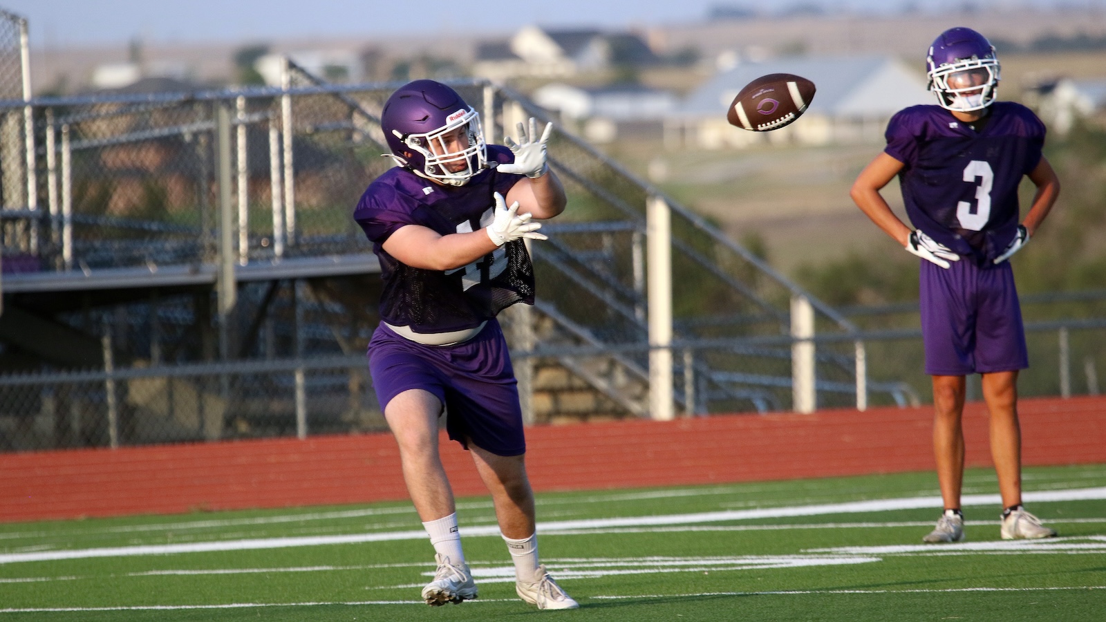 Two football players in purple uniforms on a practice field; one dives to catch a pass while the other watches from the side.