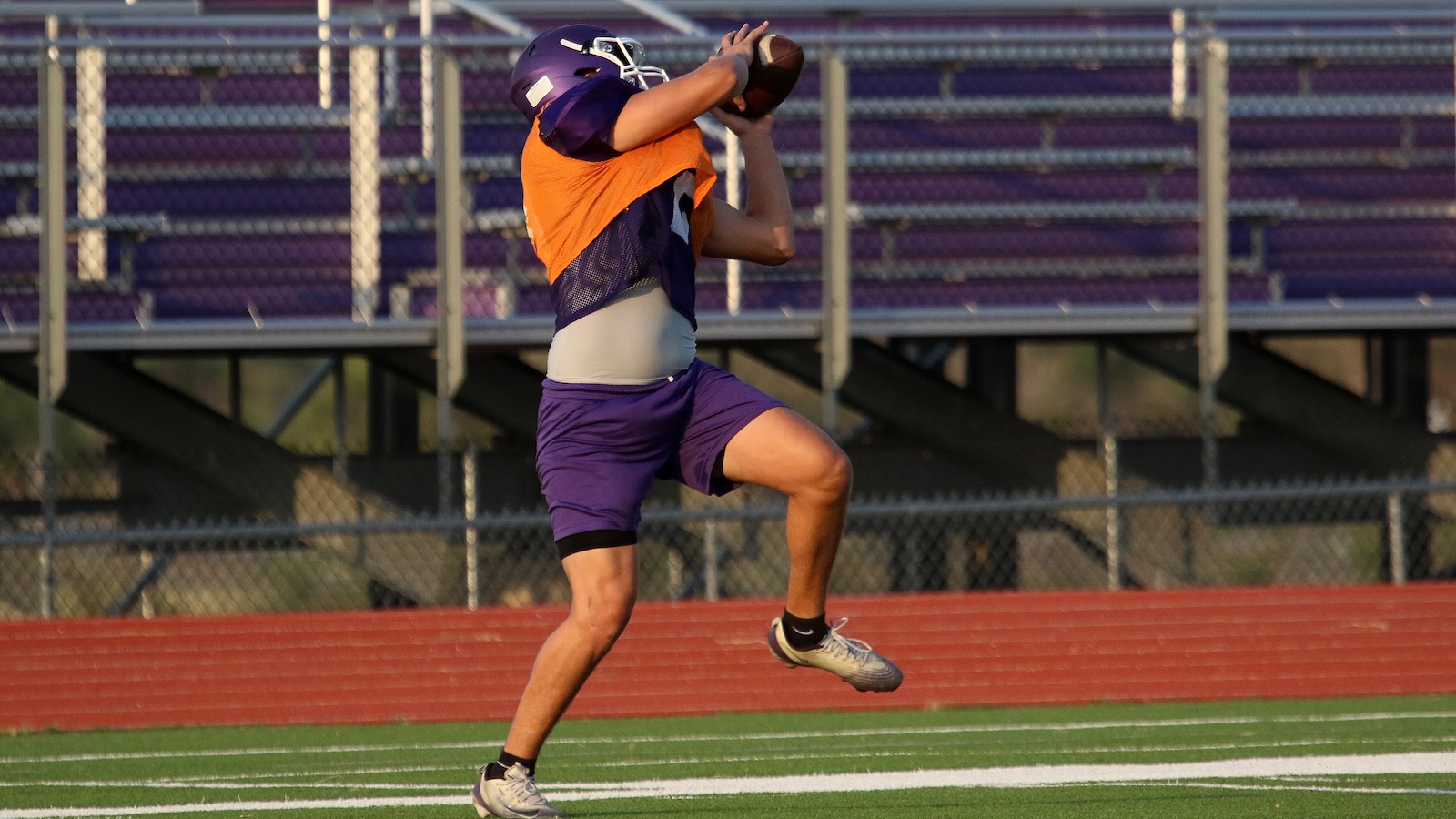 Football player in purple uniform and orange vest leaps to catch a football on a track field with empty bleachers in the background.
