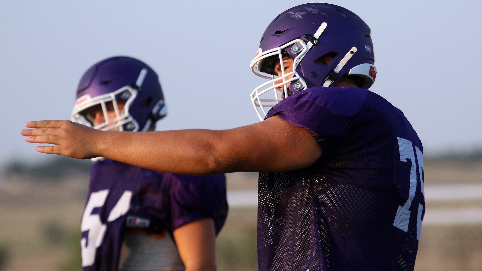 Two football players in purple uniforms practice on an open field, with one extending his arm forward as a drill.