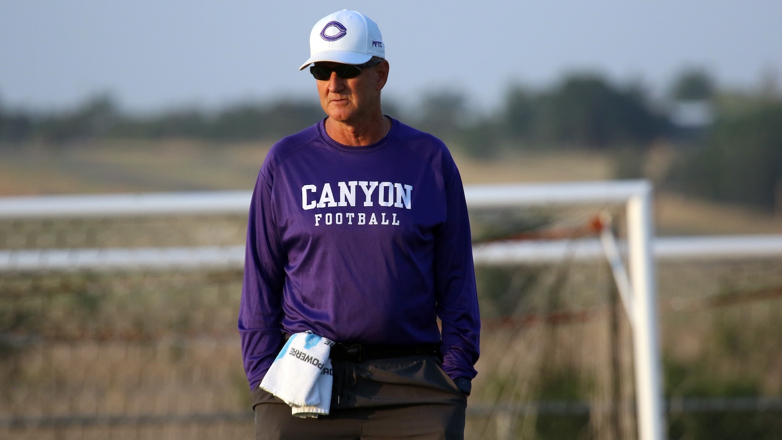 Older man in a purple Canyon Football shirt and white cap stands near a goal on a sunny training field with hands in pockets.