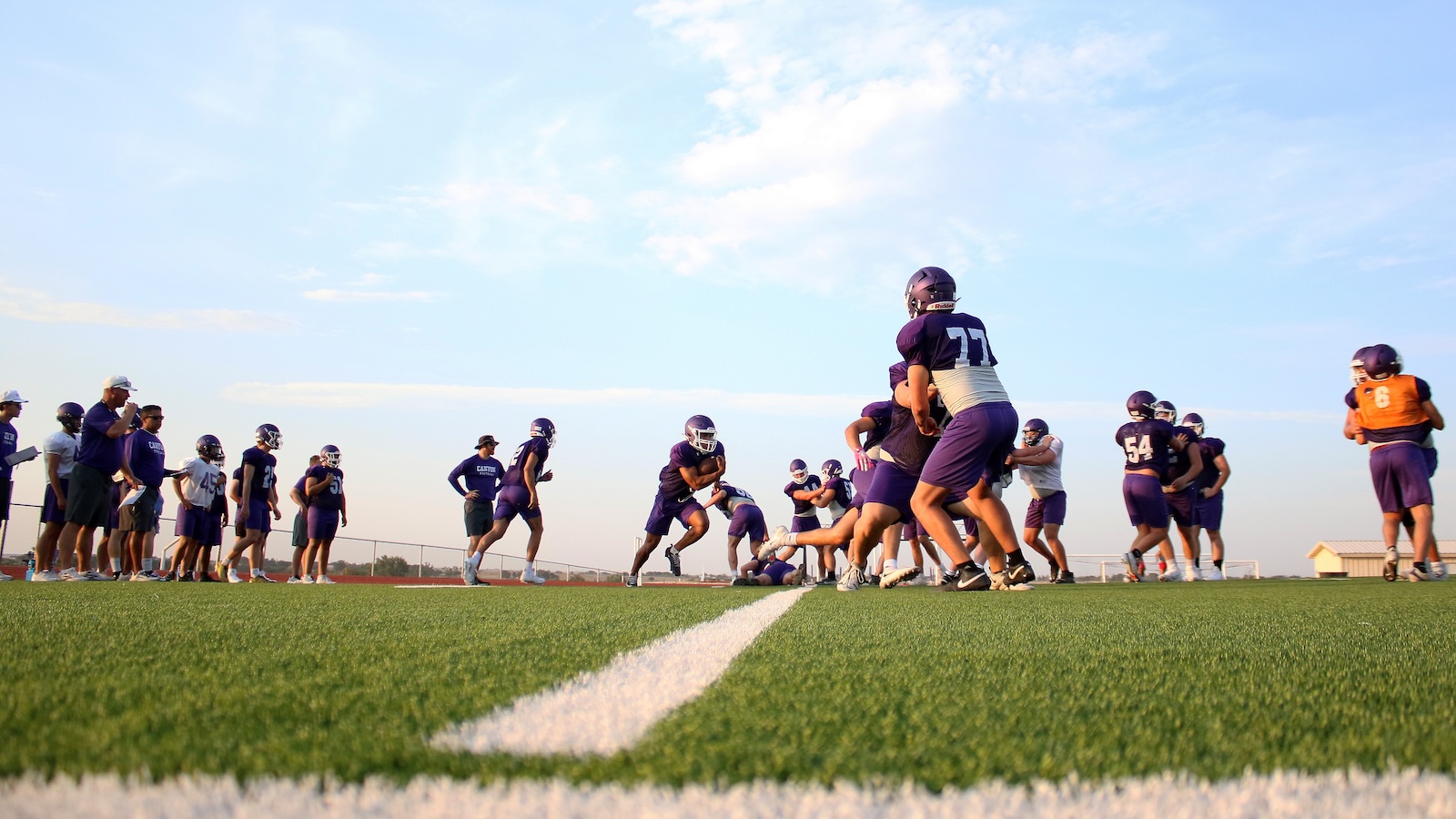 Youth football practice on a sunny field with players in purple uniforms lined up for drills on green turf.
