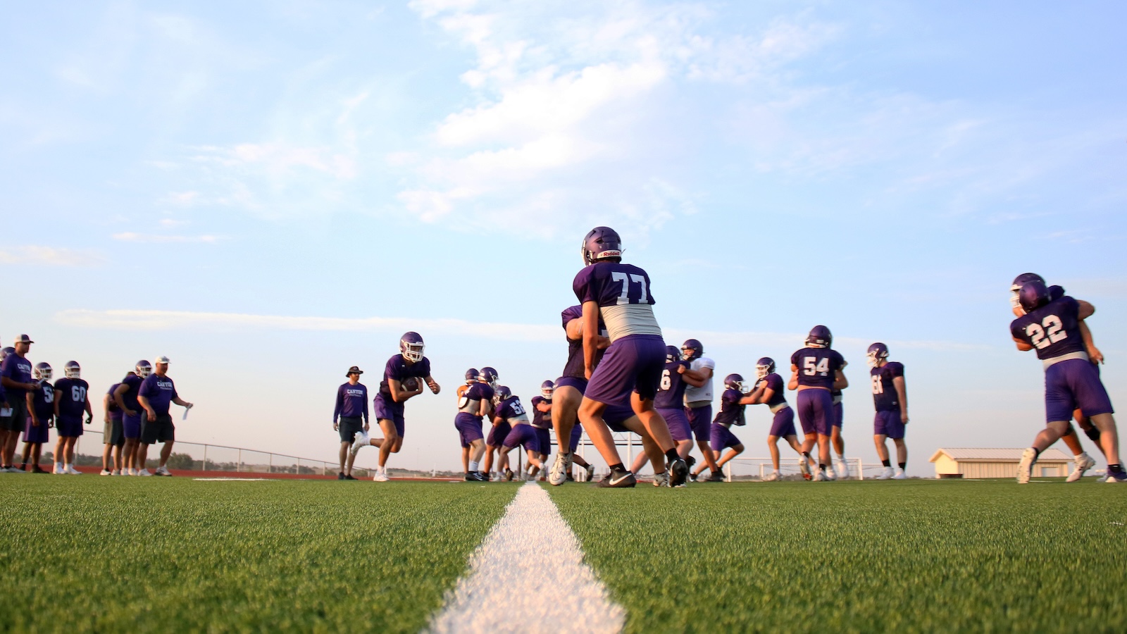 Youth football team practicing on a green field in purple uniforms under a clear sky, drills in progress.
