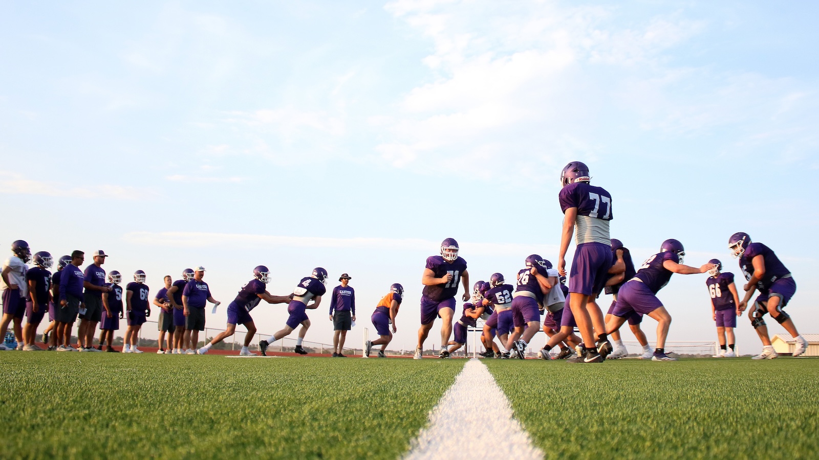 Football players in purple uniforms practice on a green field, lined up at the line of scrimmage under a clear sky.