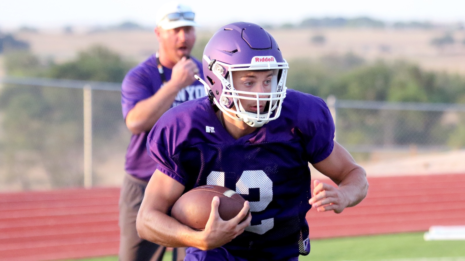 Football player in a purple jersey with number 9 runs with the ball, white helmet, during practice on a track field; a coach in purple watches in the background.