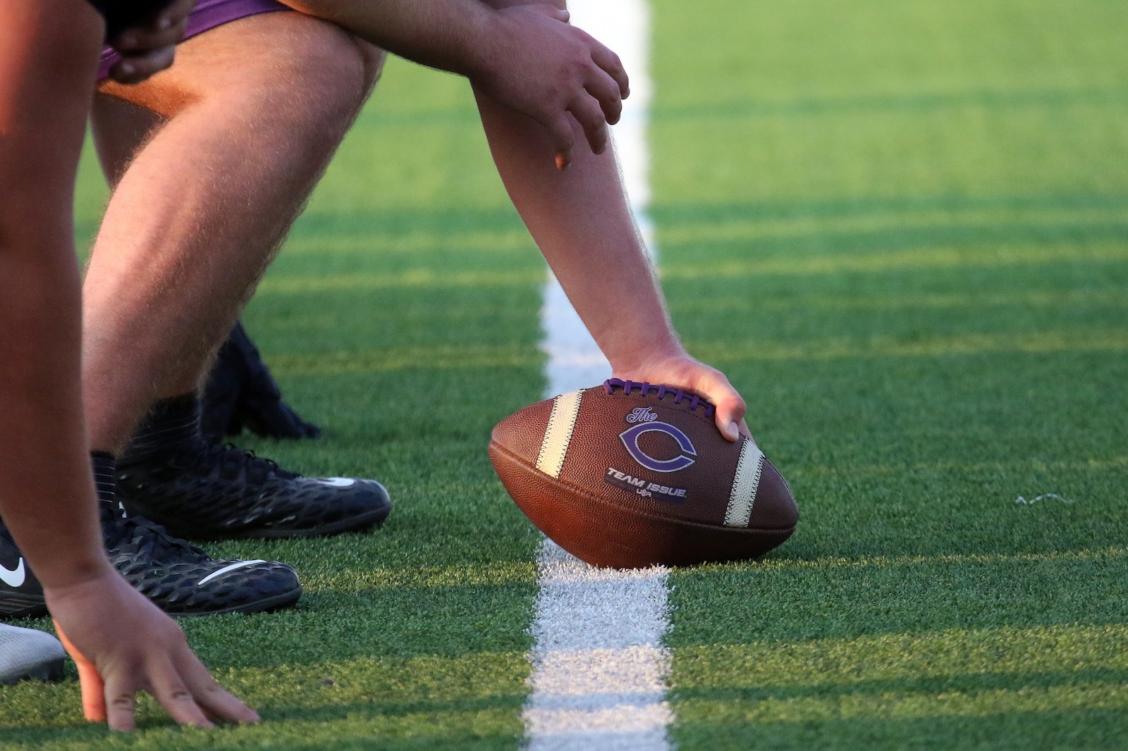 Close-up of a football resting on the line of scrimmage with players crouched behind it on a turf field.