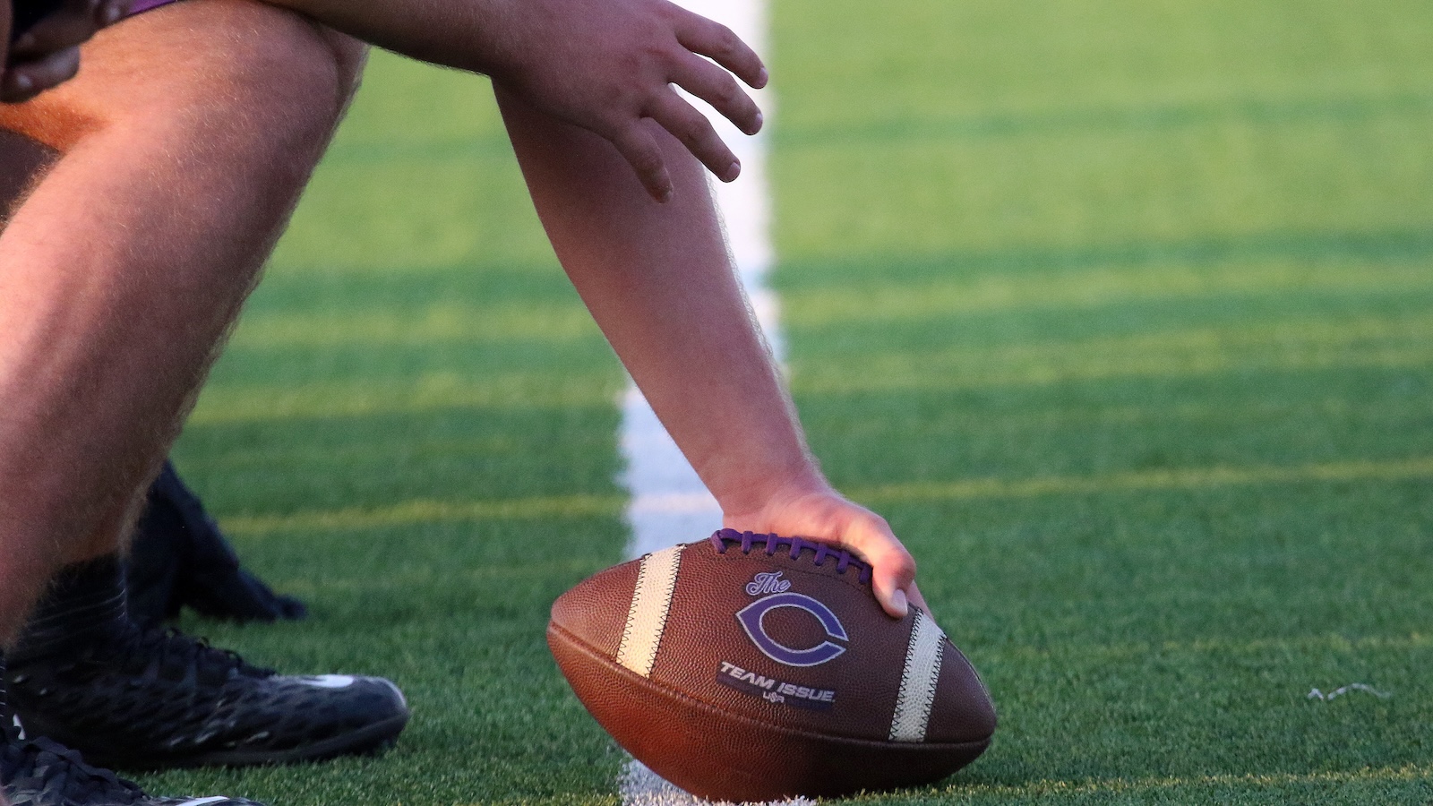 Football on a green field as a player places it for a snap, purple laces visible on the ball.