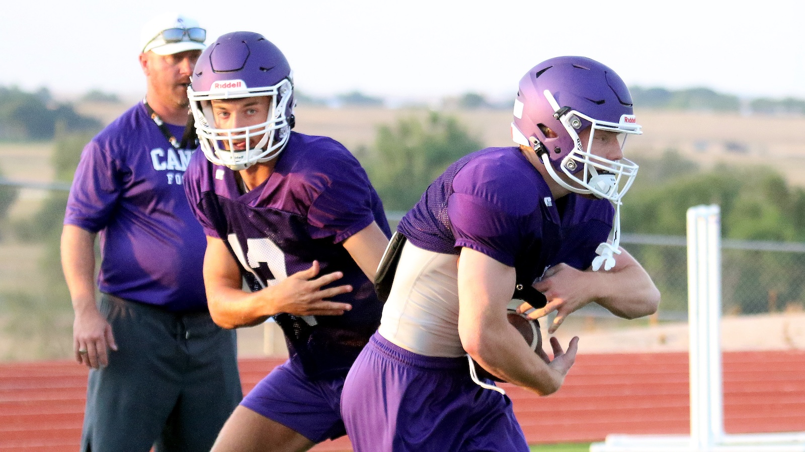 Two football players in purple uniforms on a practice field; one quarterback with the ball ready to run, another teammate nearby.