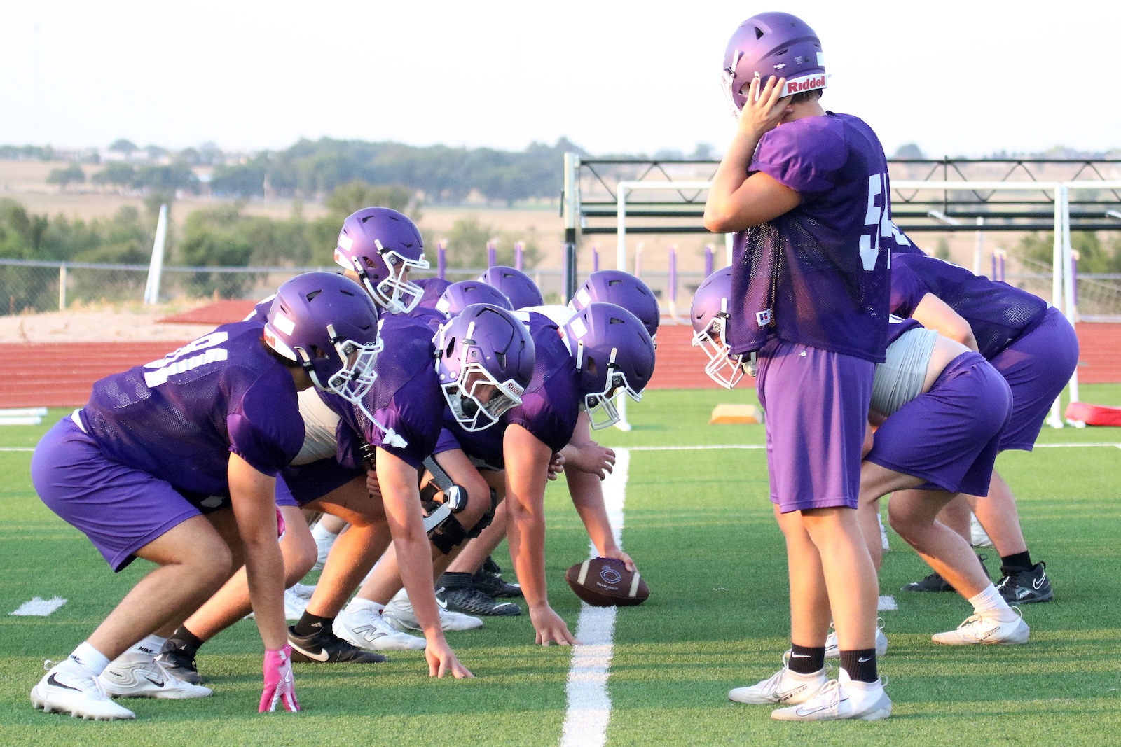 Football players in purple uniforms line up at the line of scrimmage during practice on a sunny field.