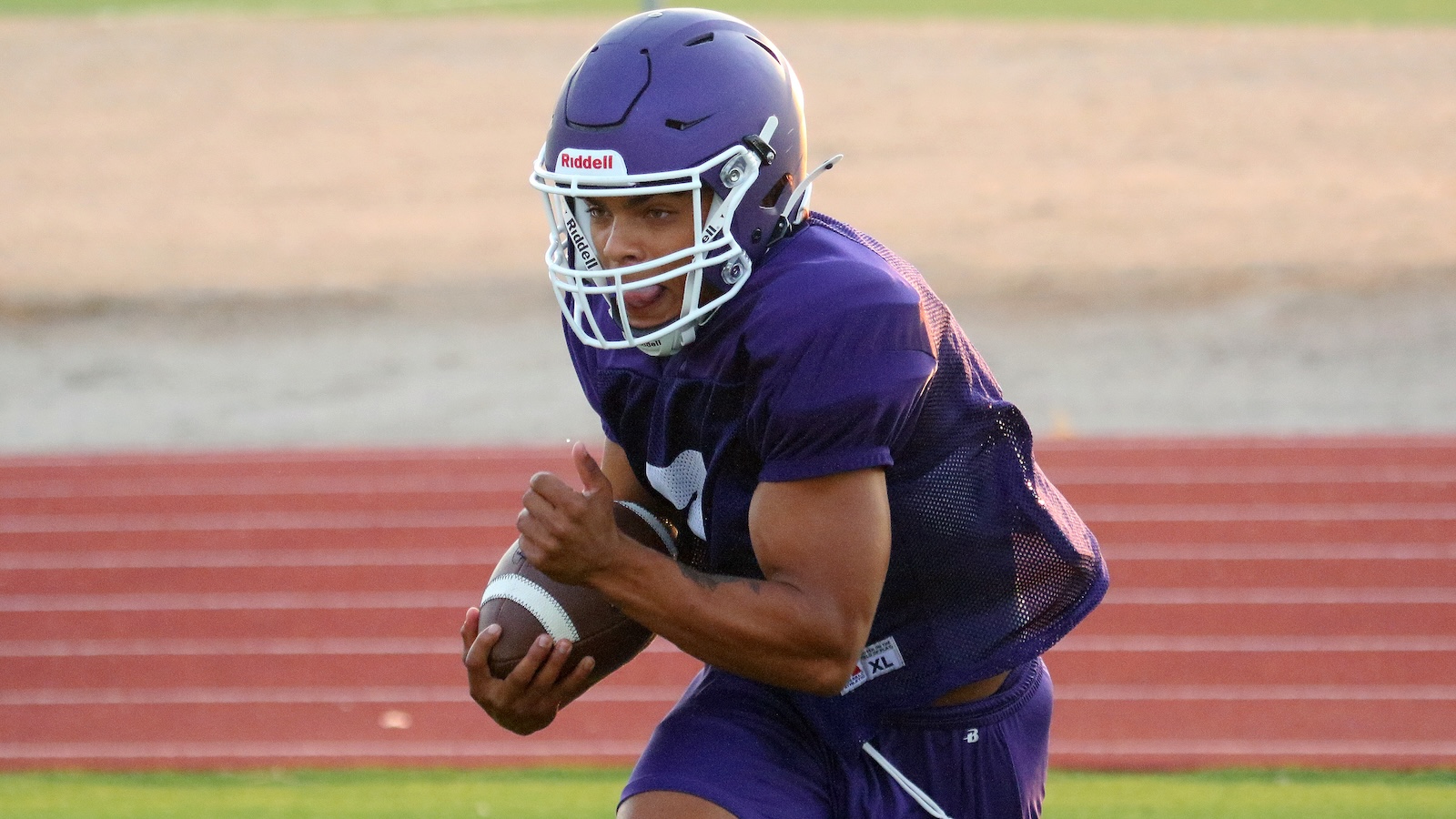 Athlete in purple football gear running with ball on a track field during practice on a sunny day
