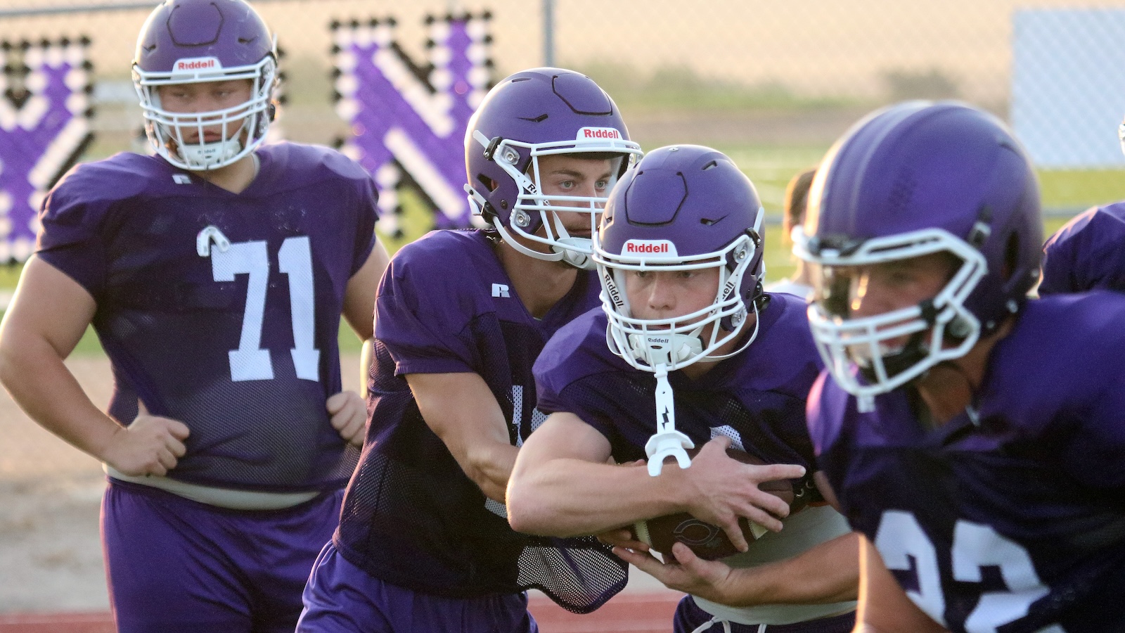 Youth football players in purple jerseys practice on a field; one player runs with the ball while teammates block
