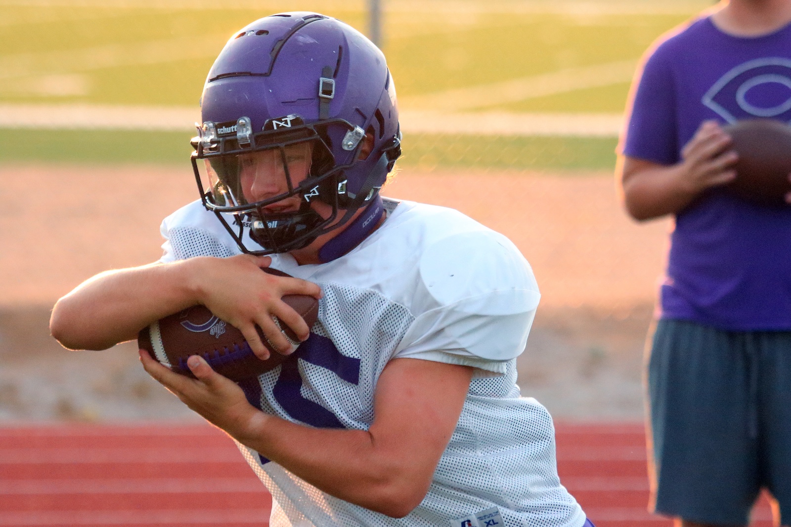 Football player in a white jersey and purple helmet grips a football, ready to run on a practice field.