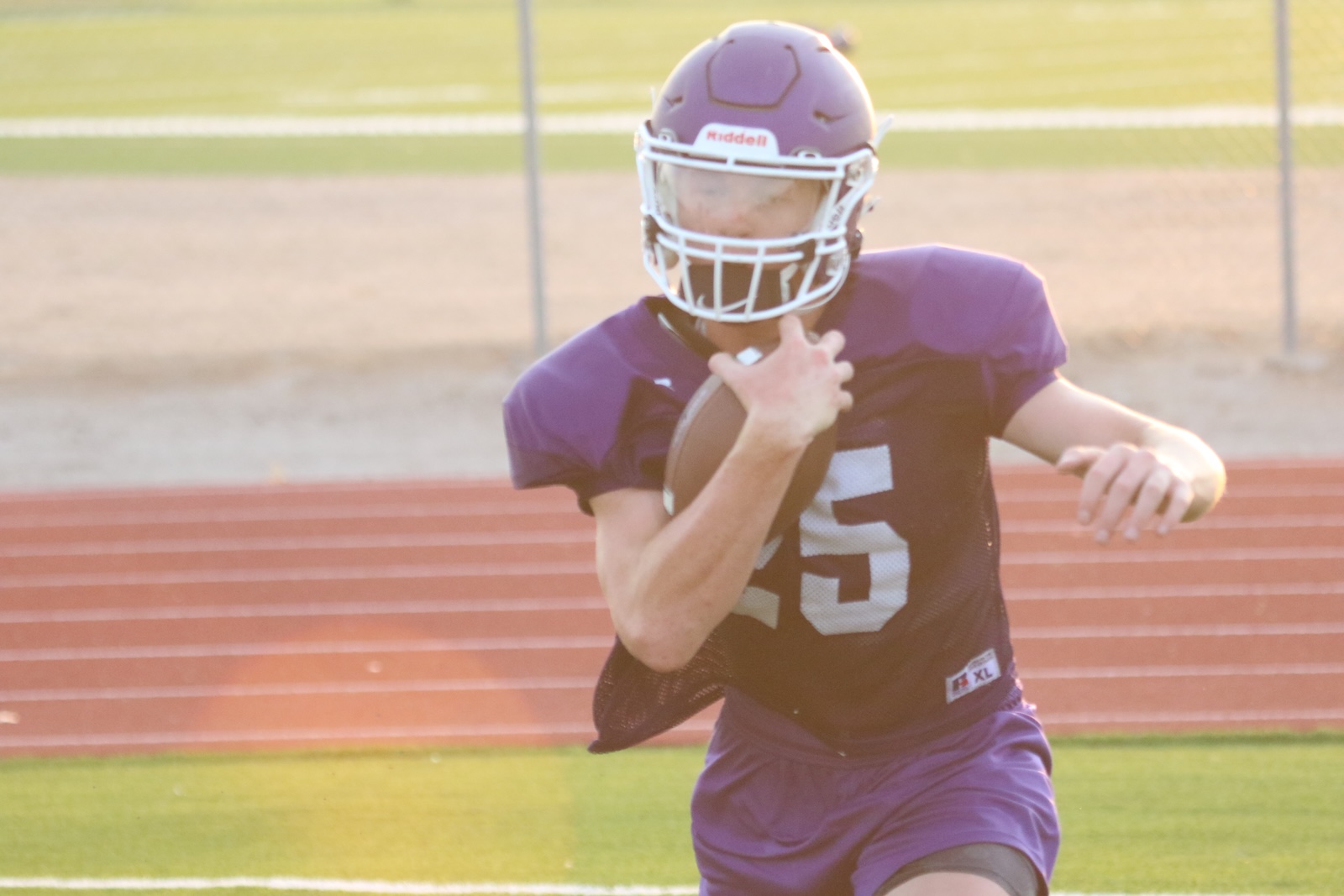 Football player in a purple jersey and helmet runs with the ball on a track field.