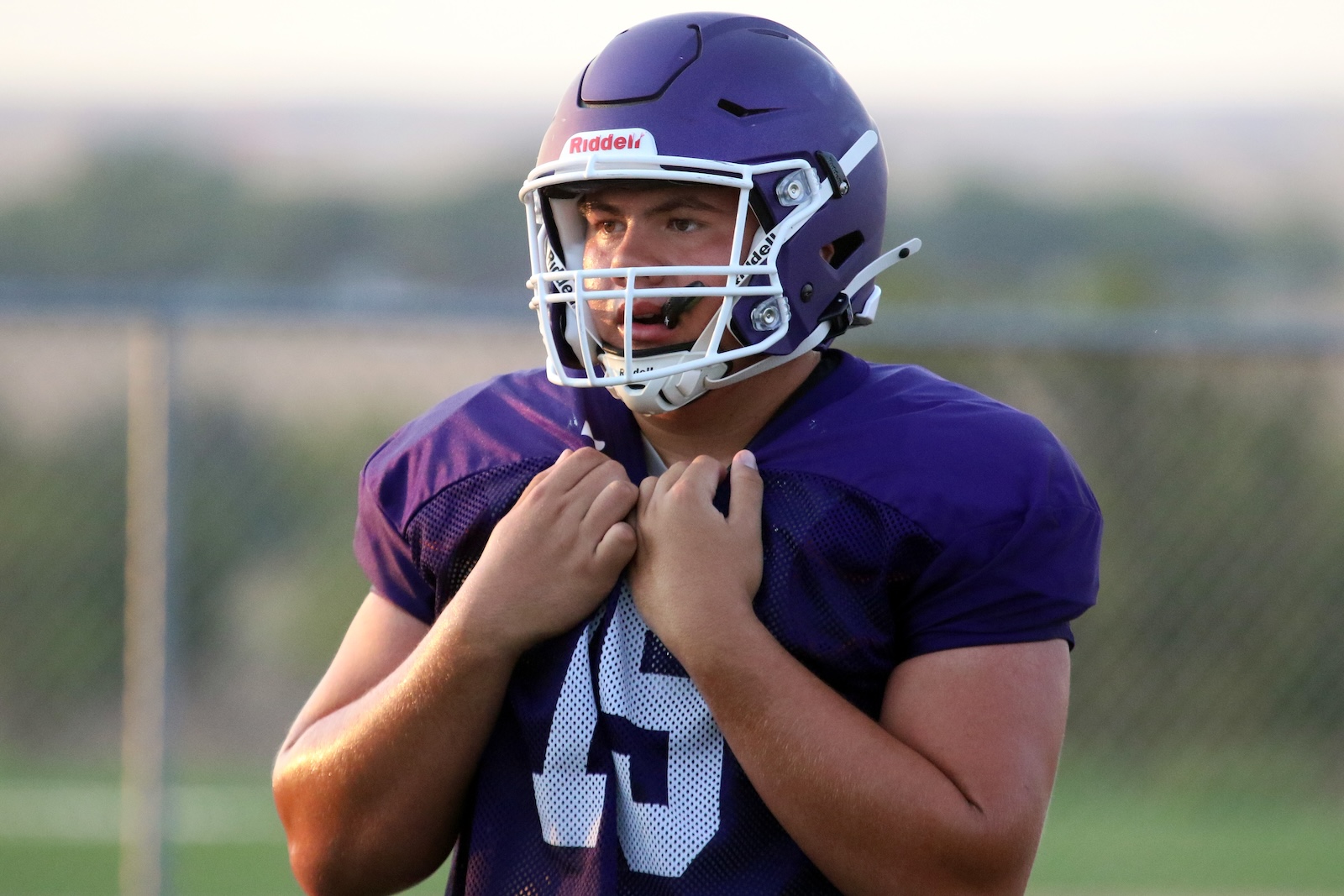 Male football player in a purple helmet and jersey adjusts his shoulder pads on a field, looking focused.