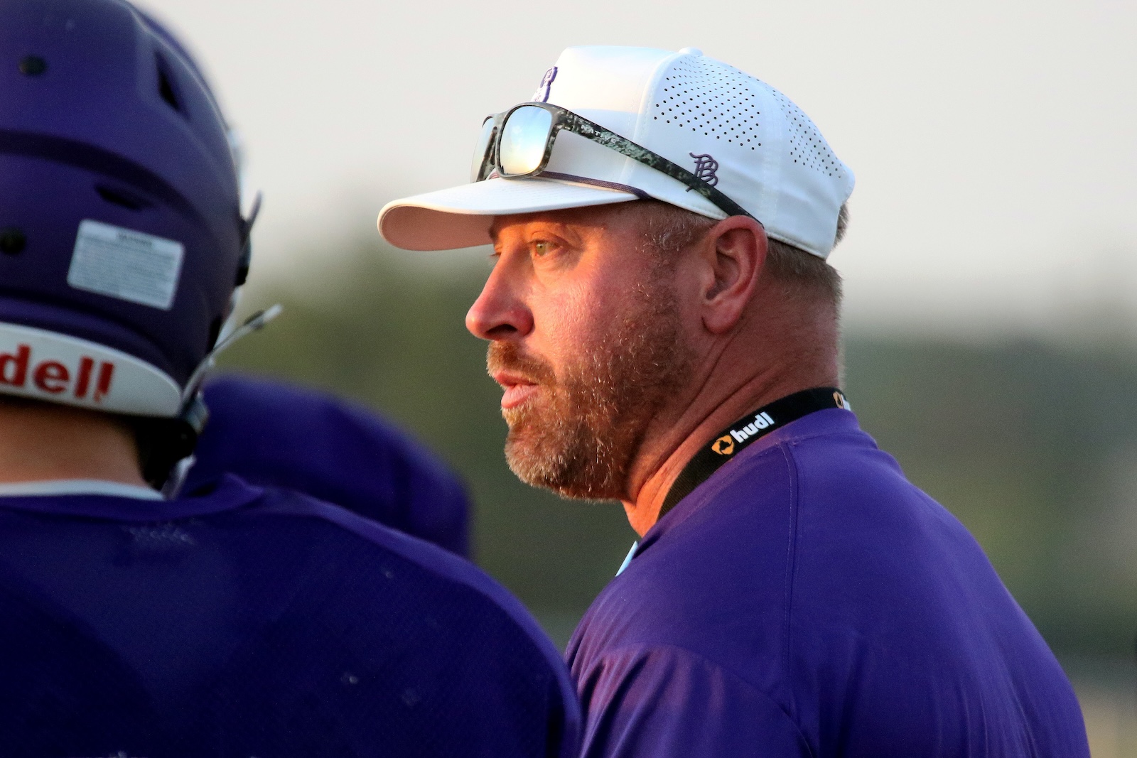 Side profile of a man in a white cap and sunglasses, wearing a purple shirt, looking intently to the right; blurred purple helmet in foreground.