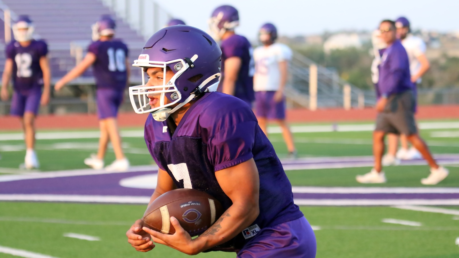 Young football player in a purple uniform runs with the ball on a practice field wearing a helmet and white facemask.