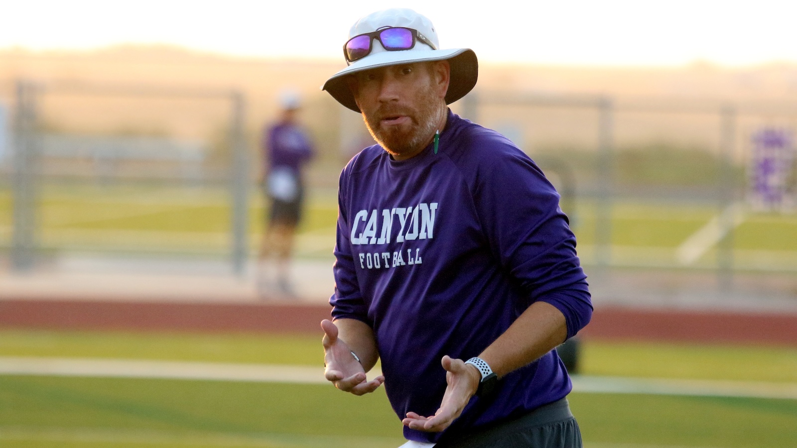 Coach wearing a purple Canyon Football shirt on a track field, gesturing with hands during practice at sunset.