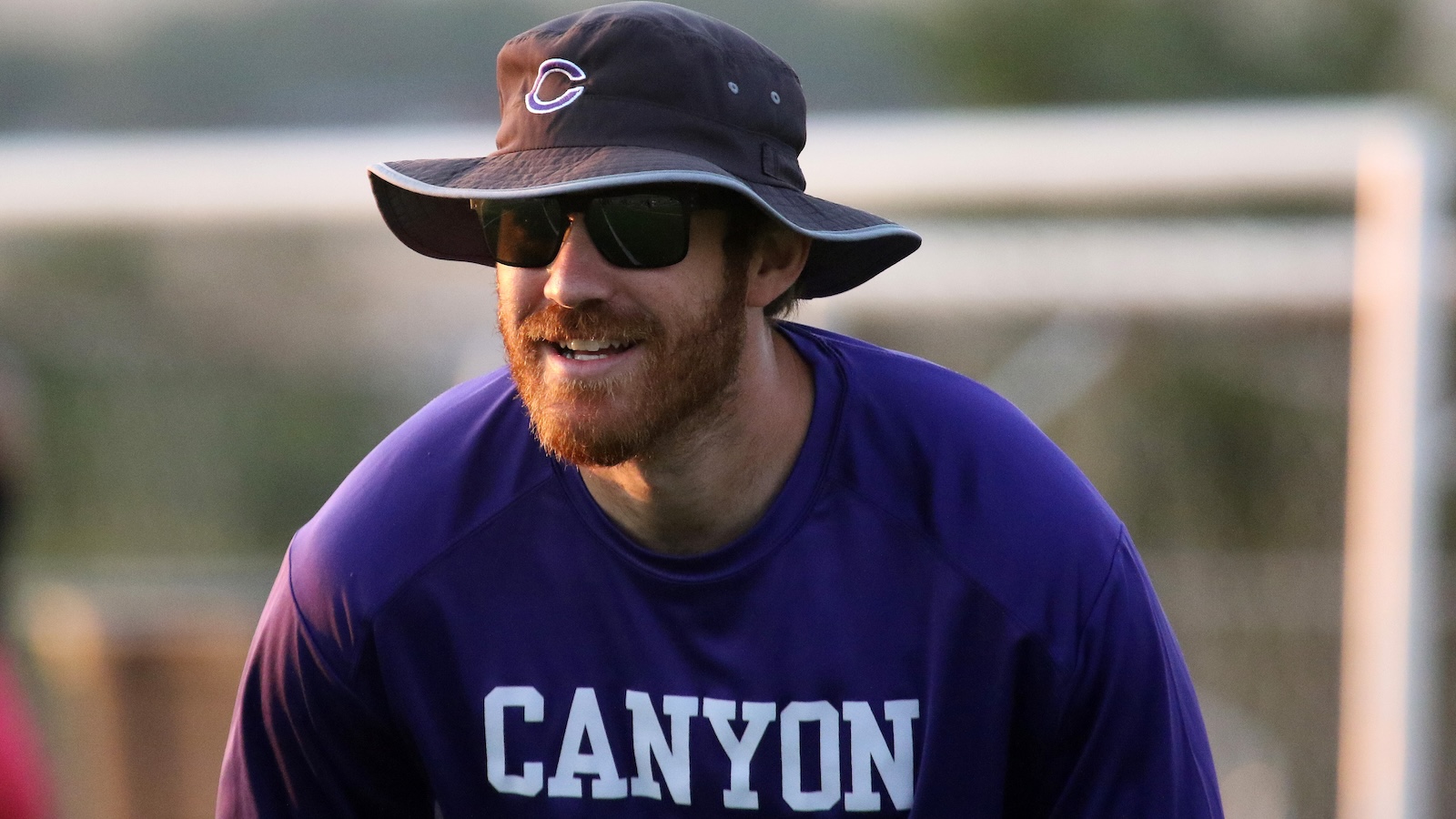 Bearded man wearing sunglasses and a wide-brim hat, smiling, in a purple Canyon shirt outdoors.