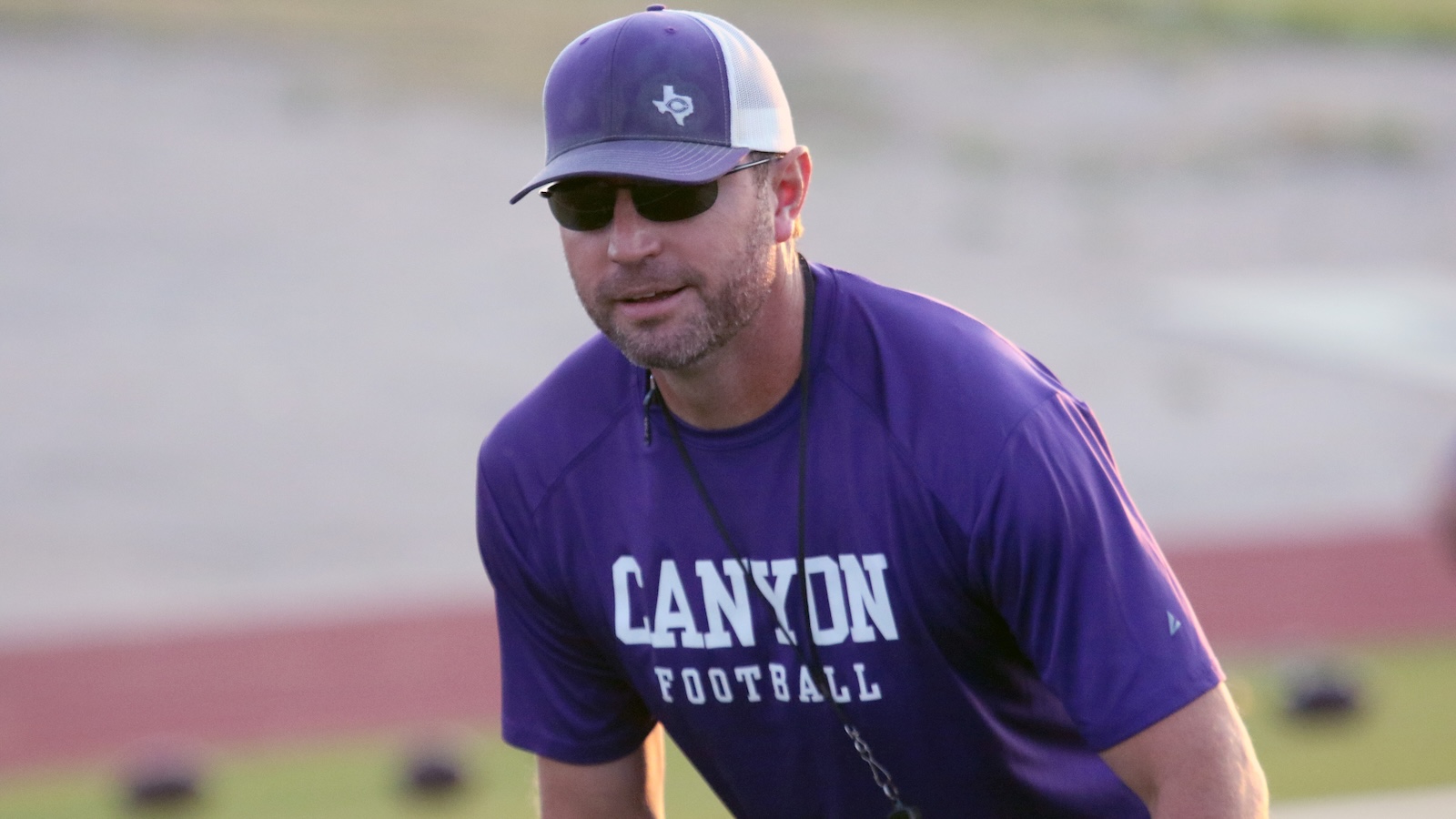 Man in a purple Canyon Football shirt, wearing a purple cap and sunglasses, coaching on a track field.