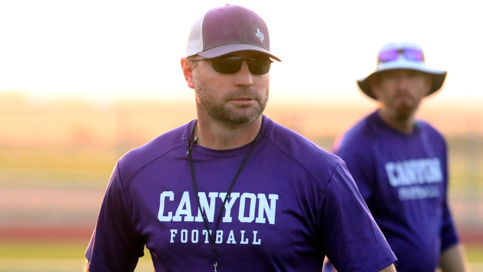 Man in a purple Canyon Football shirt with a cap and sunglasses walks on a football field; a second person in a similar shirt is blurred in the background.