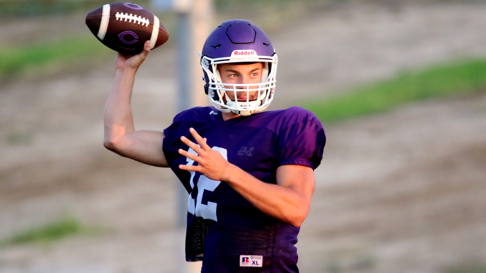 Quarterback in a purple jersey throws a football during practice, with helmet and protective facemask on a blurred field.