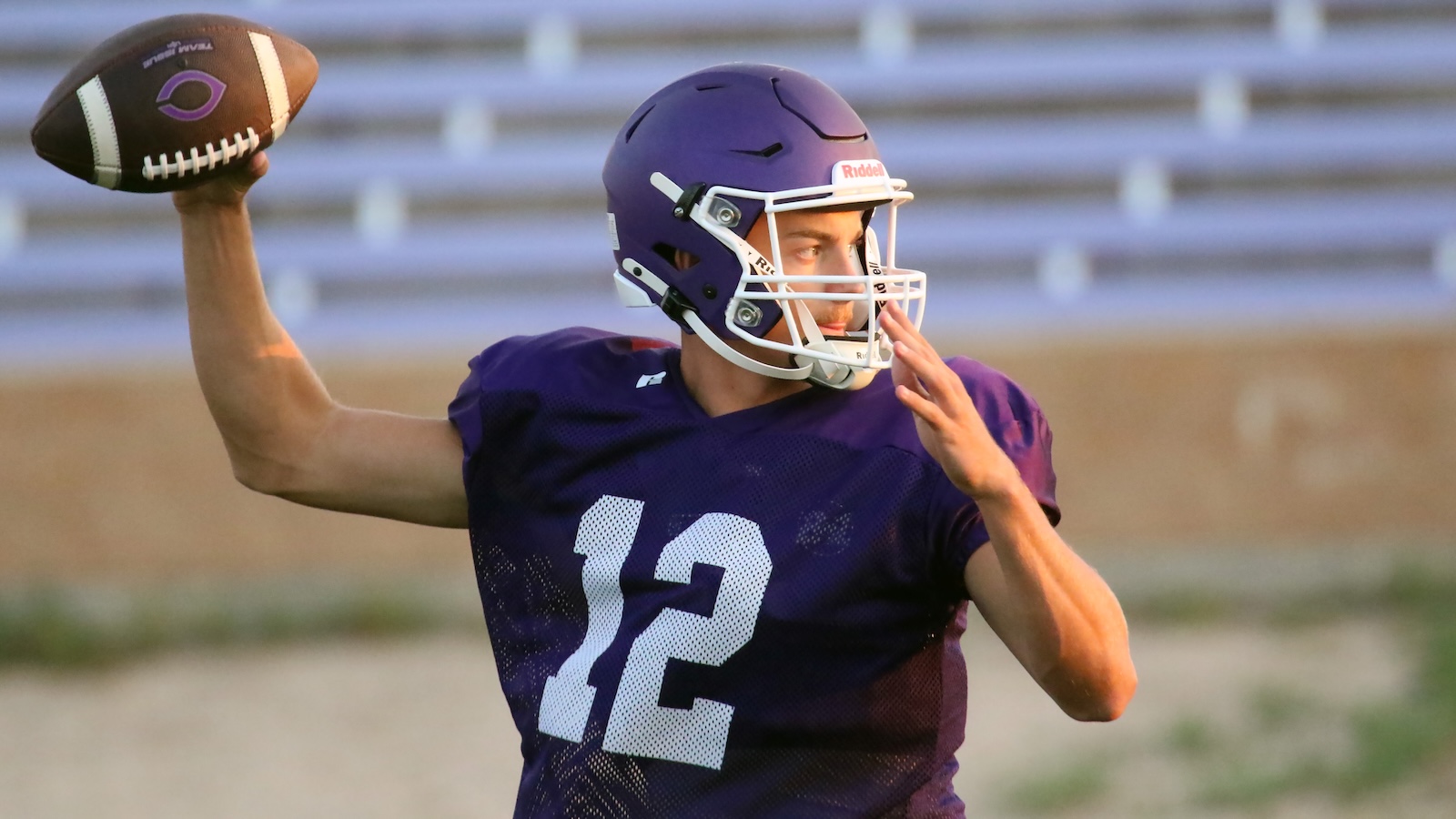 Purple-jersey quarterback in a helmet throws a football during practice with empty bleachers in the background.