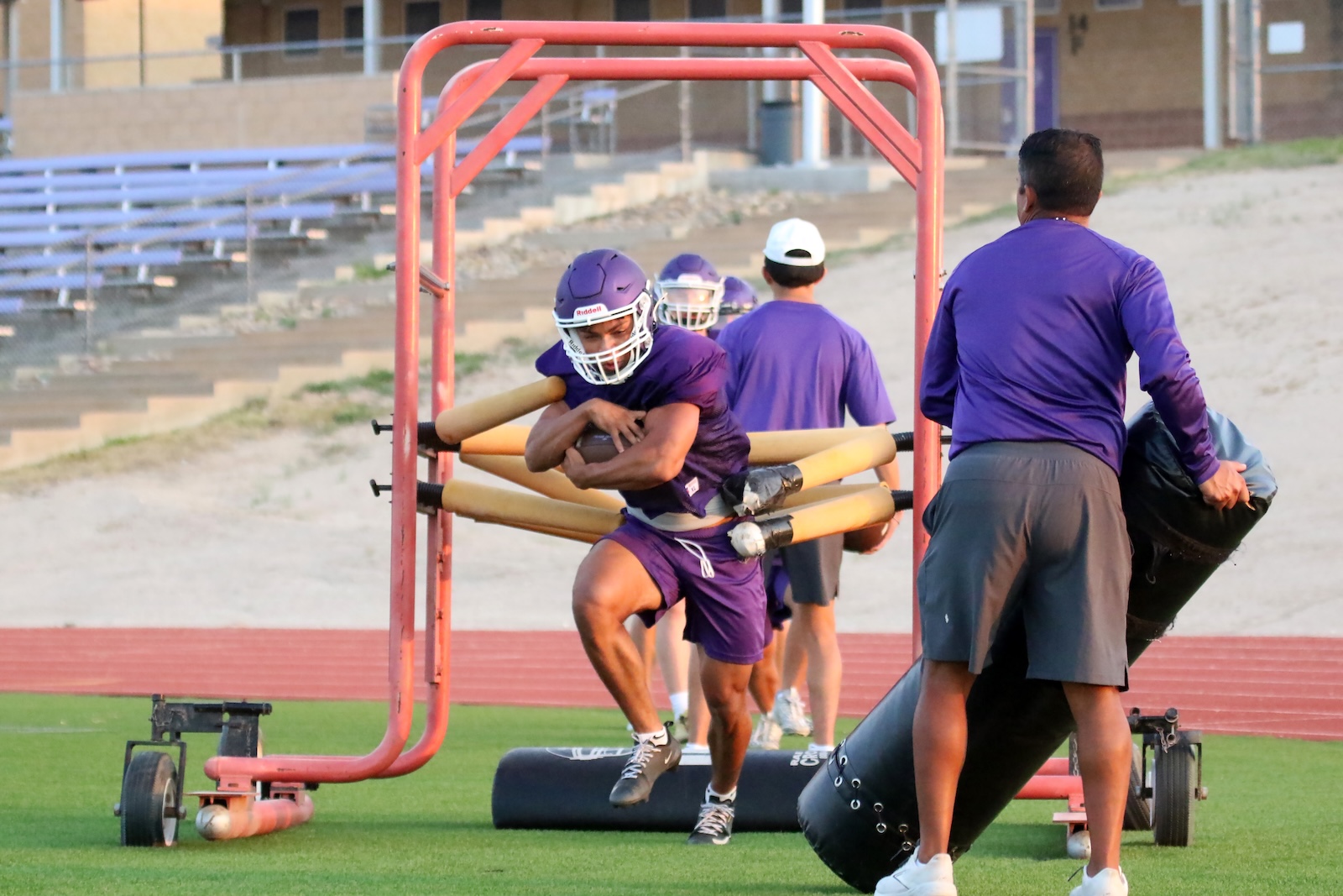 Football players in purple uniforms run a tackling drill behind a red practice sled on a grassy field.