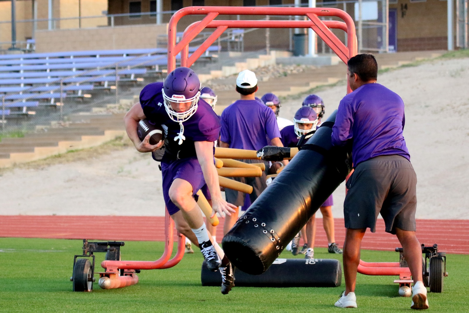 Football player in purple uniform carries ball while sprinting through hurdle frame during practice; trainer holds a large inflatable bag nearby.