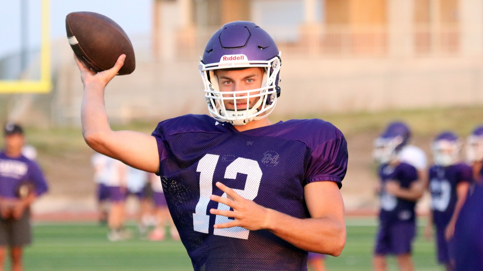 Quarterback in purple jersey (number 13) throws a football during practice on a sunny field, helmet on, focused look.