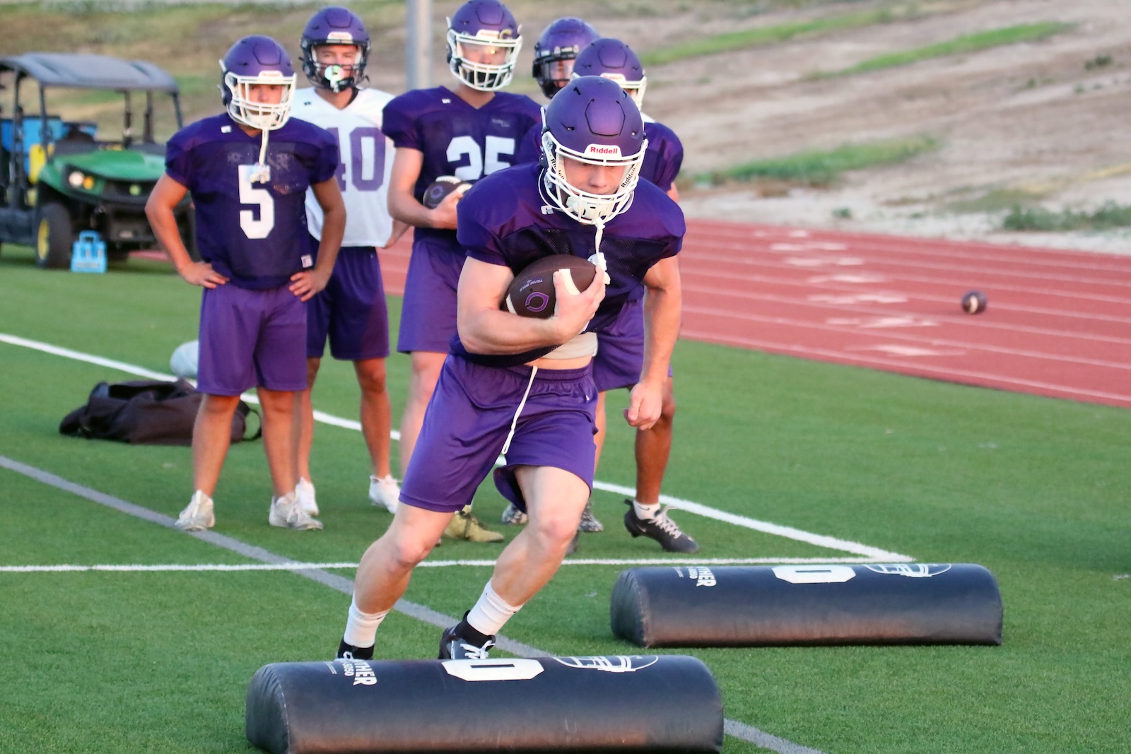 Young football player in purple uniform runs with ball during a drill on a synthetic turf field, teammates watching in the background.