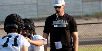Coach wearing a white cap and sunglasses blowing a whistle while guiding football players on a field