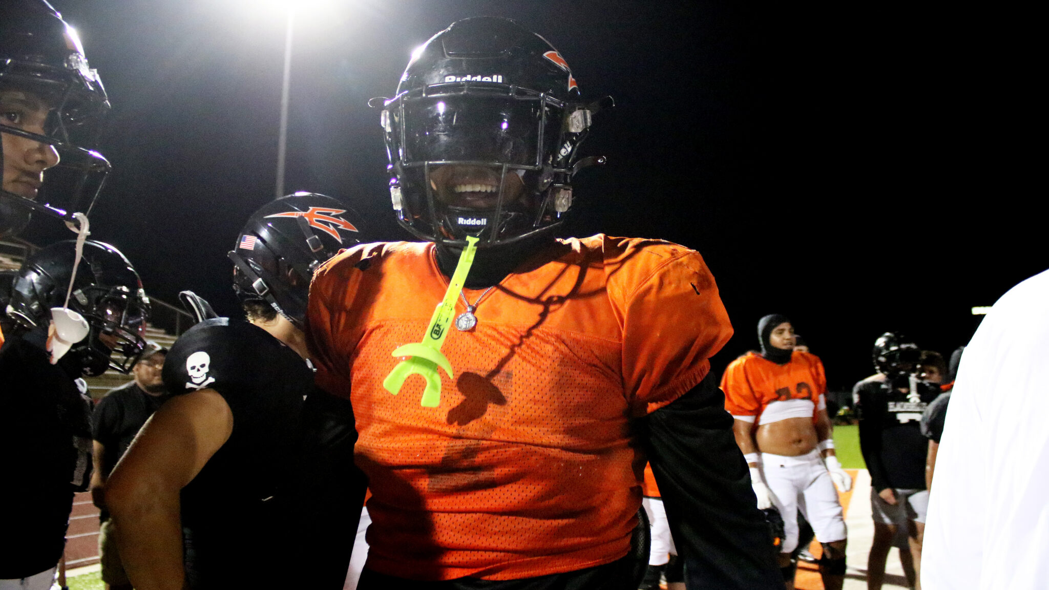 1K6A2908 | Press Pass Sports Football players in orange uniforms and helmets on a night field, one smiling toward the camera with a bright practice whistle tag visible