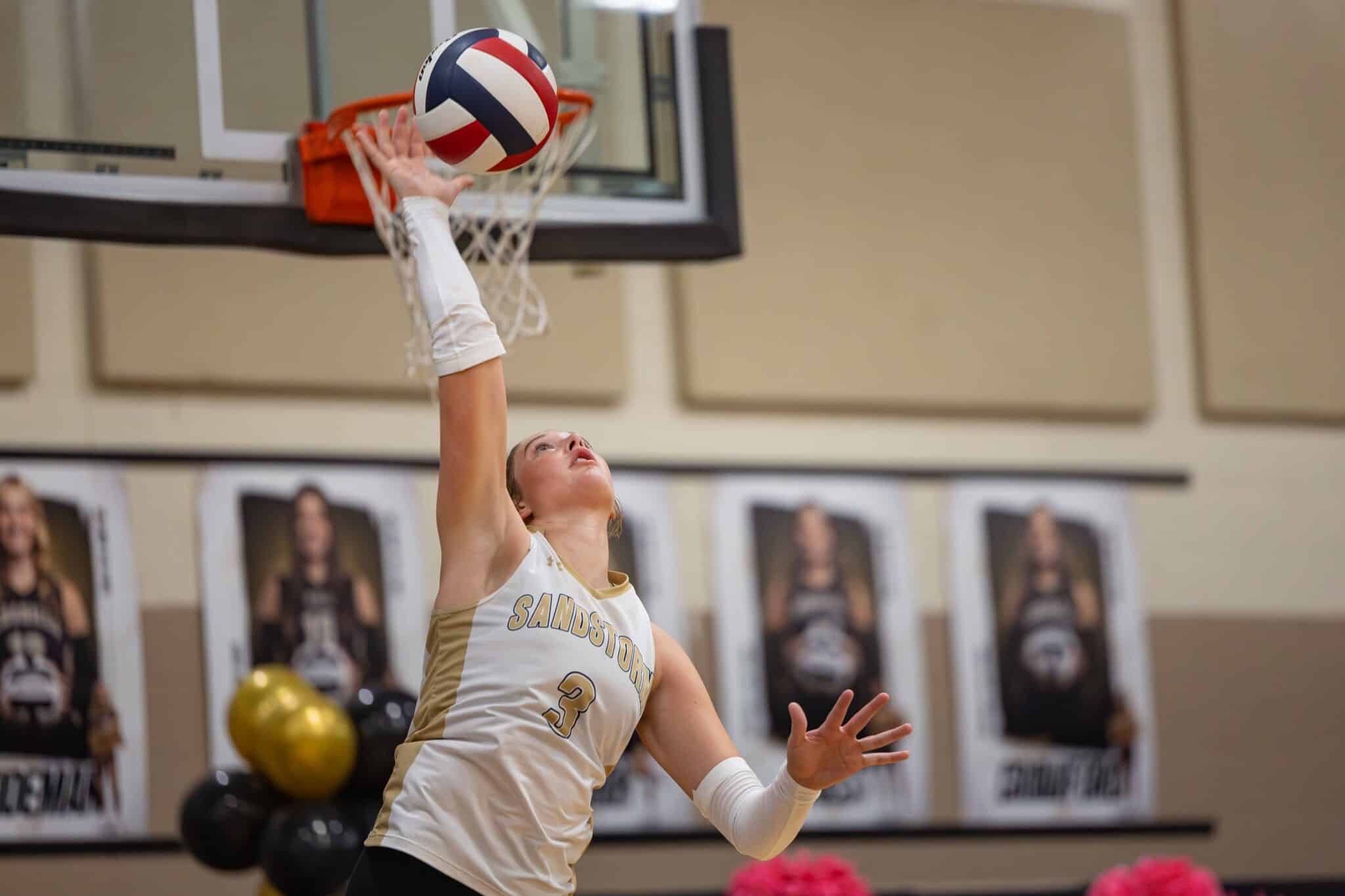 Female volleyball player in white and gold uniform reaches up to spike the ball during a game in a gym.