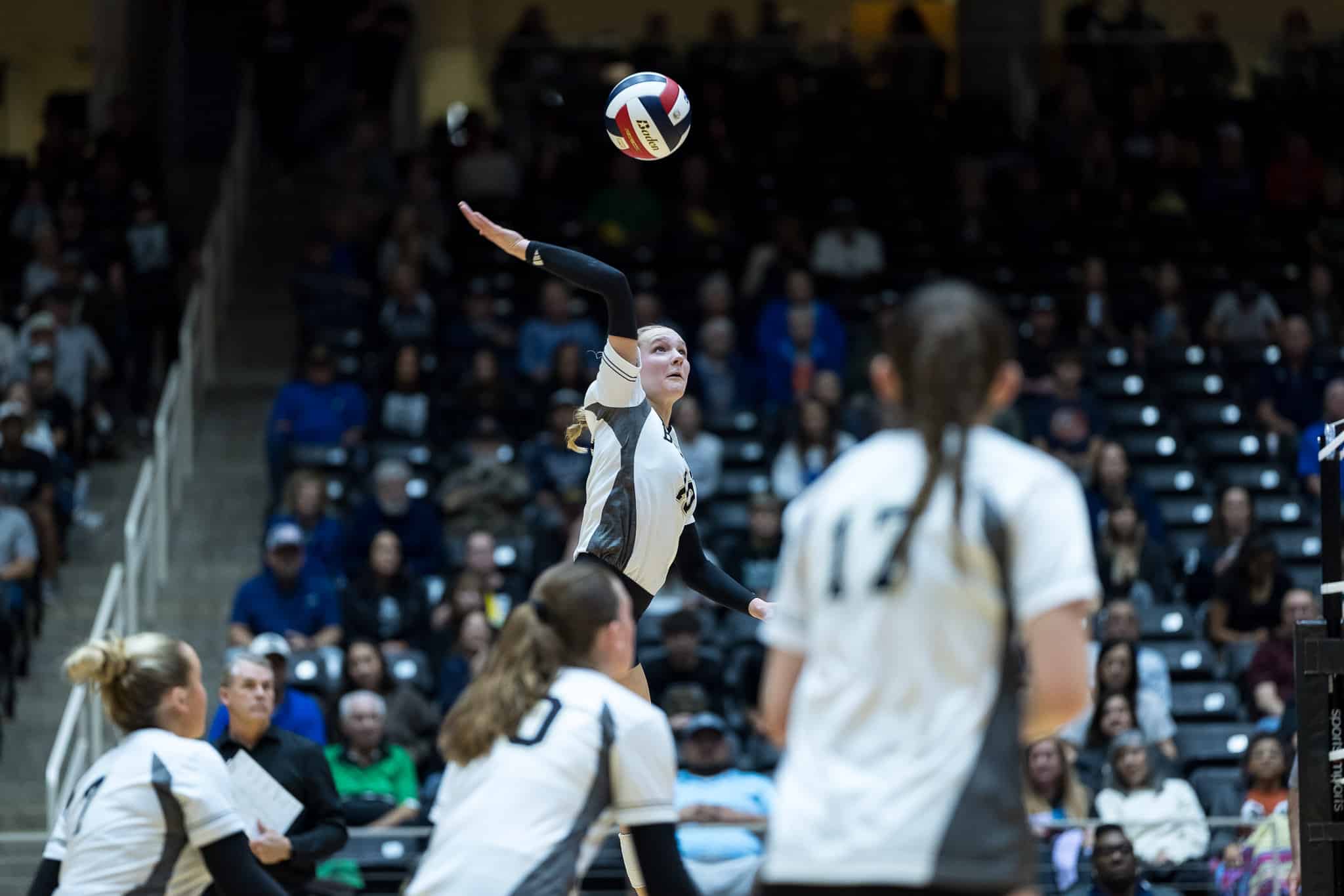 Female volleyball player in a white uniform serves the ball mid-air during a match, with spectators in the stands behind her.