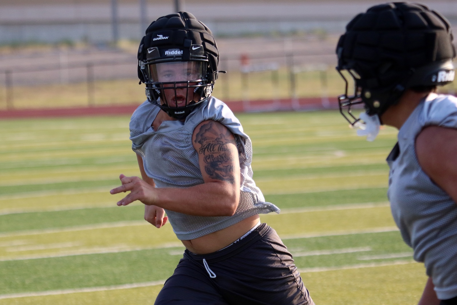 Two football players in gray practice jerseys sprint on a green artificial turf field, wearing black helmets with face guards.