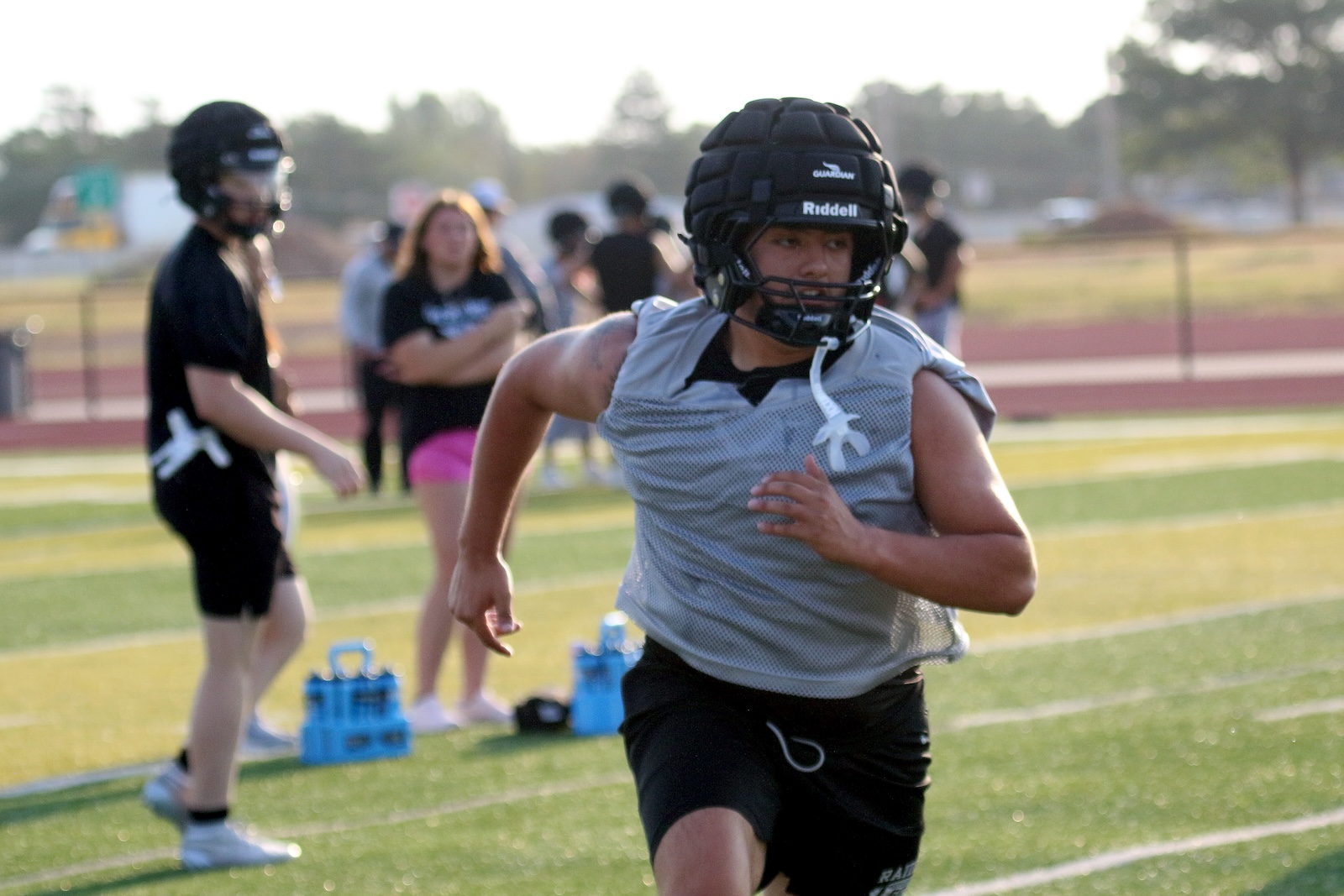 Football player in a gray practice jersey and black helmet sprinting on a turf field, with teammates and spectators blurred in the background.