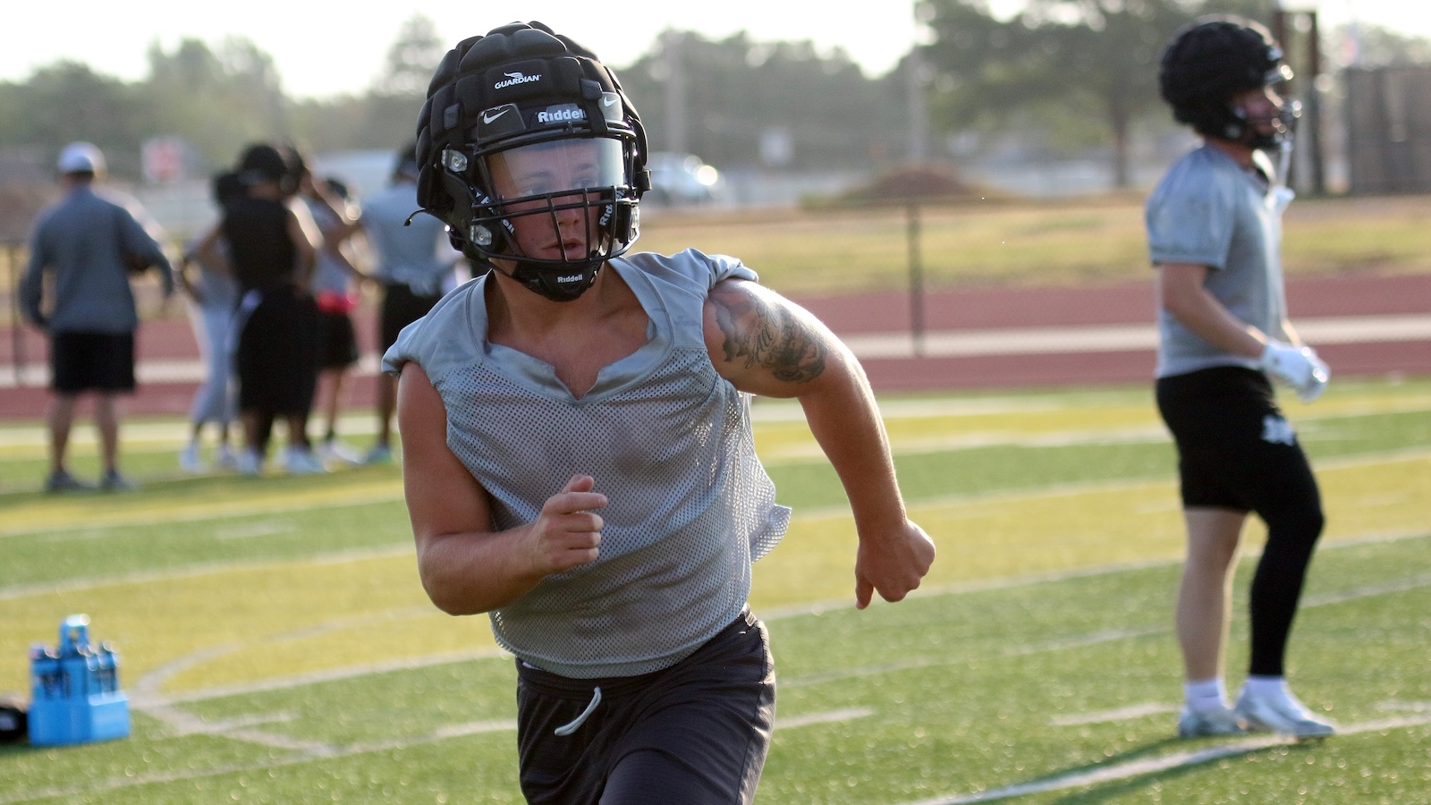 Athlete wearing a football helmet and gray sleeveless jersey sprinting on a football field, with teammates in the background.