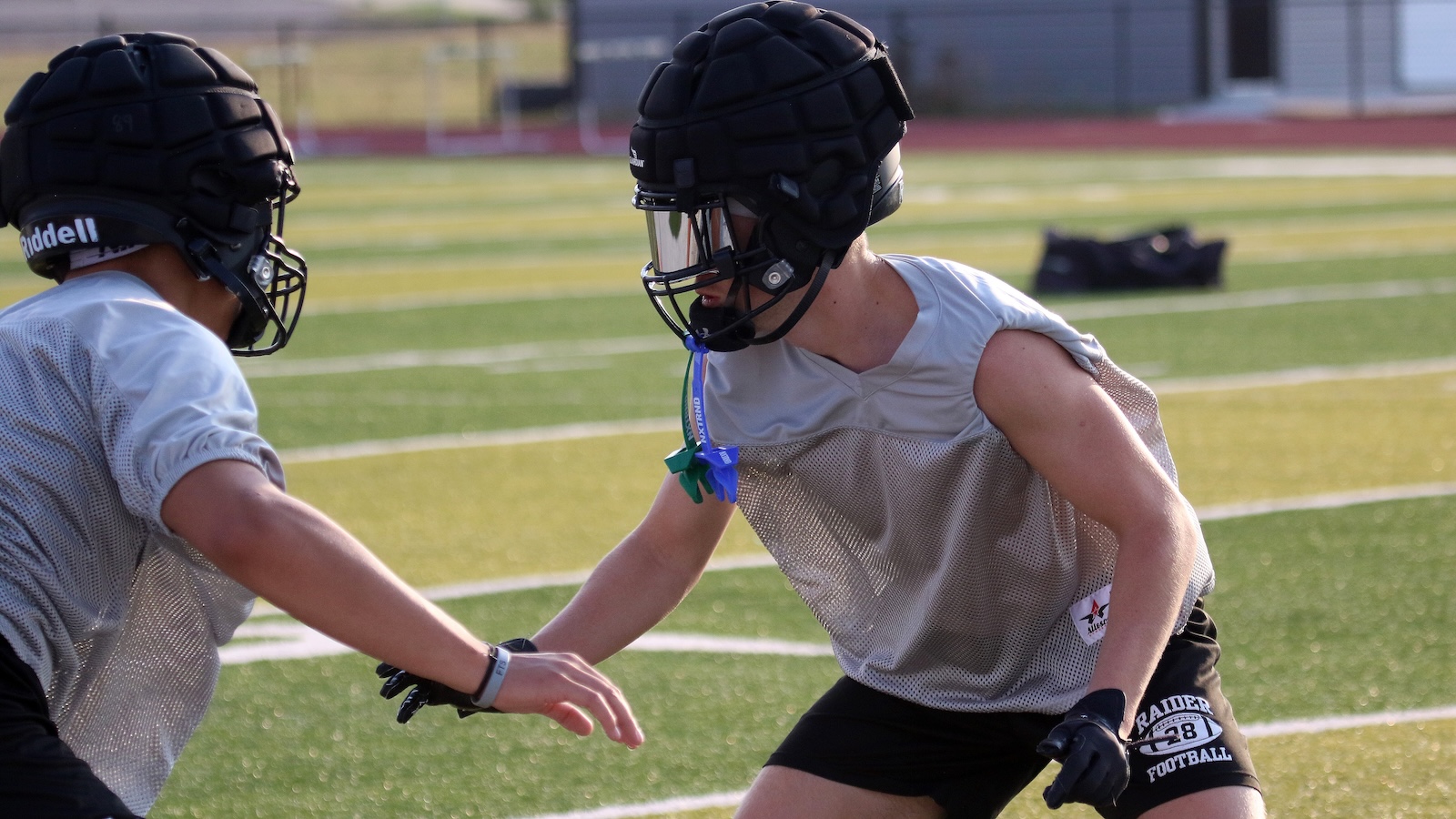 Two football players in gray mesh jerseys practice on a turf field, facing each other in helmets.